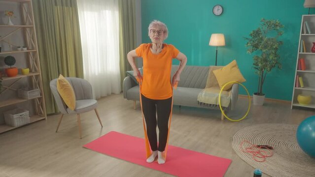 A retired woman works out in the living room with a sports elastic band. A woman, stepping on the expander with her feet, raises her hands, pulling on the elastic band. HDR BT2020 HLG Material.