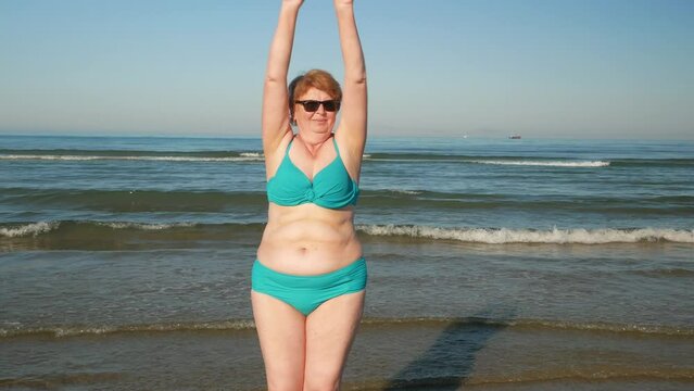 Plus size senior woman in swimsuit dancing on the sea beach in sunny day. Happy people in vacation concept. International Women's Day. Outdoors