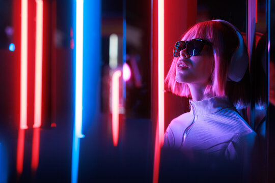 Beautiful woman with purple hair in futuristic costume over dark background. Violet neon light. Portrait of young girl in modern headphones listening music.