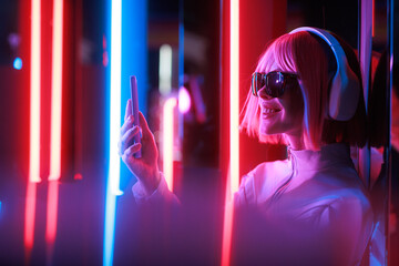 Beautiful woman with purple hair in futuristic costume over dark background. Violet neon light. Portrait of young girl in modern headphones listening music.
