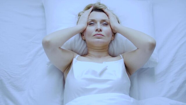 Unhappy annoyed woman massaging head lying on her bed, feeling unpleasant headache, problems with blood pressure, migraine because of menopause starting