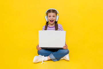 A young girl with a laptop. A little girl is sitting cross-legged on the floor with headphones on,...