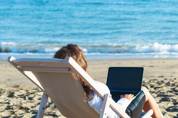 Back view of alone young brunette woman freelancer in office clothes with a laptop looking on the sea sitting on the deck chair on beach. Summer concept