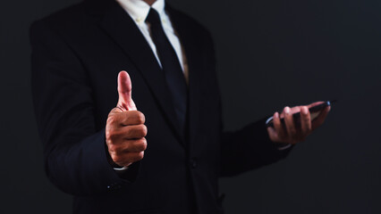 Businessman showing thumb up while using smartphone against dark background.