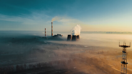 Chimneys of thermal power plant rise from fog on horizon line against sky. View of pipes from...