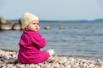 Cute baby toddler sitting on pebbles by the sea in the cold season