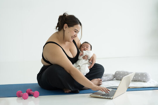 Happy smiling Asian young mother doing exercises and using laptop computer on yoga mat during taking care her newborn at home