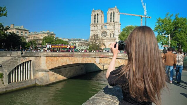 A young woman takes a picture of the Gothic-style Notre Dame Cathedral. A fashionable girl walks to see the Catholic cathedral with flying buttresses designed for the church in Paris, France.