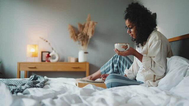 Concentrated curly african woman in eyeglasses reading book with cup of tea on the bed at home
