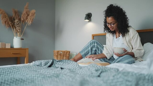 Lovely curly african woman in eyeglasses reading book with cup of tea on the bed at home