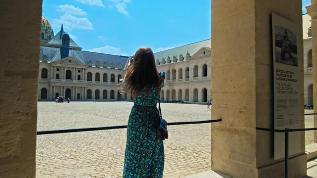 A young woman visiting and taking pictures of The Court of Honor of Hotel National des Invalides in Paris. A tourist looking at the Golden Dome of the Hotel des Invalides and rooftops of Paris.