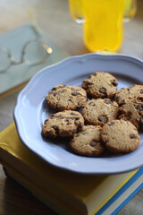 Plate of chocolate chip cookies, stack of books, reading glasses, orange soda, mobile phone and pen on the table. Hygge at home. Pastel colors, selective focus.