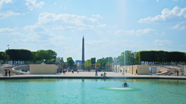 Tourists taking walks in Tuileries Garden in Paris, France. People relax in a large public park looking at the beautiful green lake with the Place de la Concorde in the background.