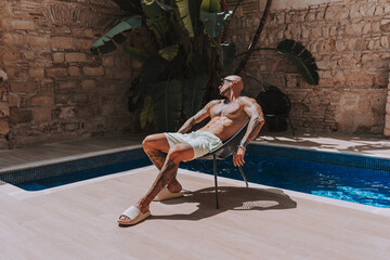 Fototapeta na wymiar Chico joven musculado y tatuado posando sin camiseta en piscina de estilo boho en un hotel un dia soleado de verano