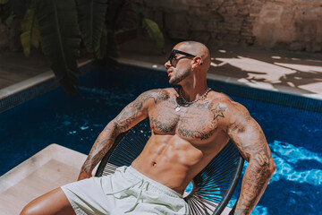 Chico joven musculado y tatuado posando  sin camiseta en piscina de estilo  boho en un hotel un dia...