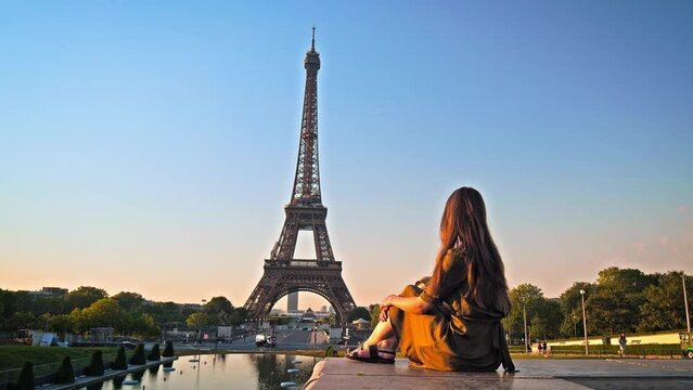 A beautiful woman in a dress sitting at a fountain and looking at the Tower of Paris, France. A female tourist meditates on the beauty of Paris at the golden sunset.