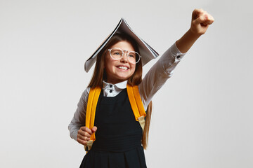 Funny young schoolgirl acting like superhero, holding open book on head and raising up fist while...