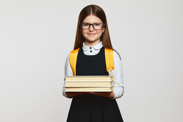 Smart happy school girl wearing yellow backpack and eyeglasses, smiling for camera and carrying bunch of books against white background.