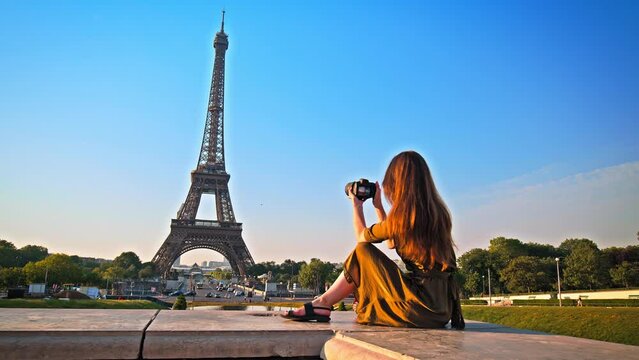 A beautiful woman in a dress uses a camera to picture the romantic city of Paris, France. Female tourist taking pictures of the historical Tower and enjoying the view of a golden sunset.