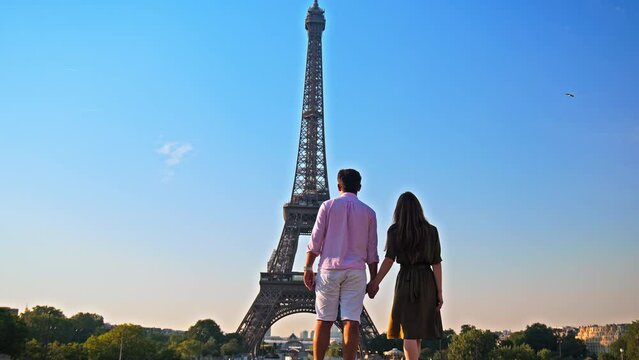 A young couple enjoys the view of Paris pointing at the Tower. A romantic woman and her boyfriend walk together holding hands at golden sunset in Paris, France.