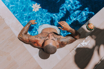 Chico joven musculado y tatuado posando  sin camiseta en piscina de estilo  boho en un hotel un dia soleado de verano