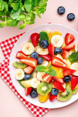 Summer fruit and berry salad with fresh strawberries, blueberries, banana, kiwi, orange and mint, pink background, top view