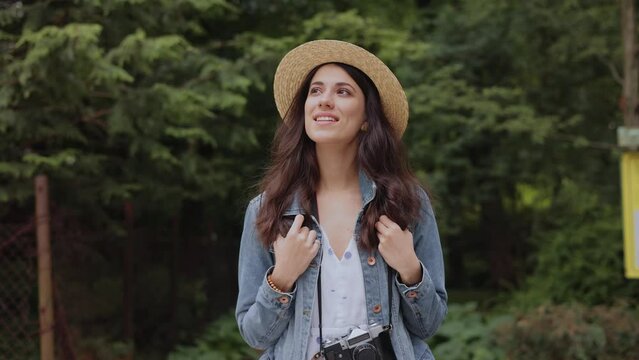 Portrait of young tourist girl in hat with beautiful eyes walking and looking around the park. Happy Woman in hat with camera enjoing time outdoor. Hipster traveller woman in hat walking in forest.