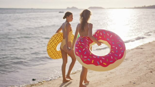 Two girlfriends walking along sunset beach holding hands after swimming in sea with pineapple and flamingo inflatables. Women on vacation and weekend fun