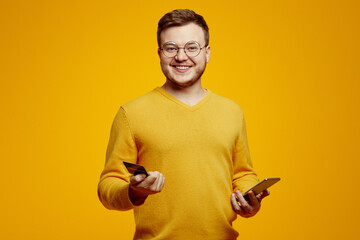 Young caucasian overjoyed bearded man wears orange sweater and glasses, holds smartphone and credit...