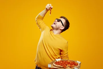 Tuinposter Excited man with sunglasses holding and biting tasty slice of pizza, posing with open mouth, holding cardboard flat box on yellow orange background © Davidovici