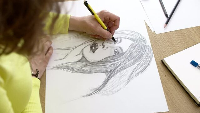 The artist draws a pencil portrait of a young beautiful woman. The female creates a picture of a woman with a dark pencil on white paper.