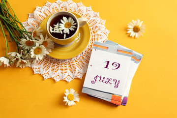 Calendar for July 19: name of the month July in English, numbers 19, a cup of tea on an openwork napkin with chamomile, a bouquet of daisies next to it, yellow background