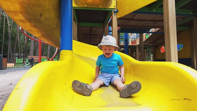 A small child is crying on a slide. The boy does not want to leave the playground. Summer family walk. Copy space - the concept of fear, falling, pain, prohibitions, resentment, whims and tantrums