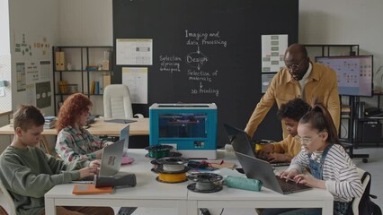 African American teacher walking around classroom and helping kids with coding on laptops during 3D printing lesson in primary school - Powered by Adobe