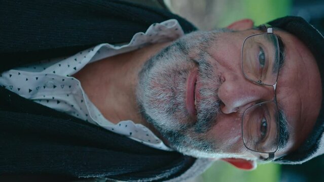 Senior Hispanic man with gray beard in eyeglasses posing for the camera outdoors in the park. Vertical format footage, video portrait
