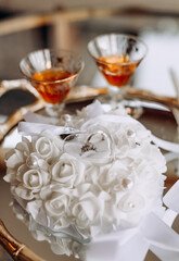 Wedding rings on a white pillow and two glasses of cognac on a tray. Wedding accessory in the form of gold rings and alcohol. Wedding day and rings for the bride and groom close-up