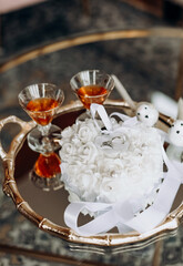 Wedding rings on a white pillow and two glasses of cognac on a tray. Wedding accessory in the form of gold rings and alcohol. Wedding day and rings for the bride and groom close-up on a vertical image