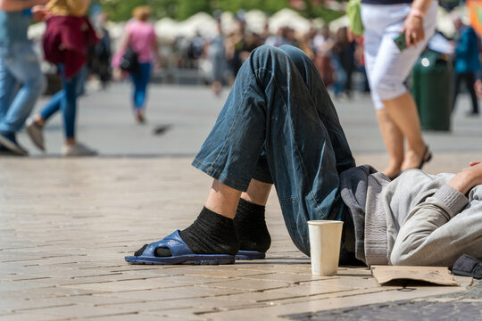Authentic tramp sleeping on a cobbled street on a sunny summer day with a paper cup for donations against the background of passers-by and tourists. Global connectivity, diversity and inclusion