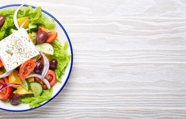 Traditional Greek Salad with Feta Cheese, Tomatoes, Bell Pepper, Cucumbers, Olives, Herbs in white ceramic bowl on White rustic wooden table background from above, Cuisine of Greece. Copy space