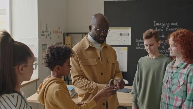 Medium shot of group of primary school students learning 3D printer details with African American teacher during computer class