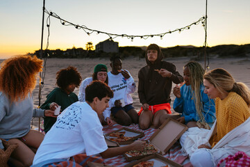 Happy diverse friends eating pizza on beach at evening