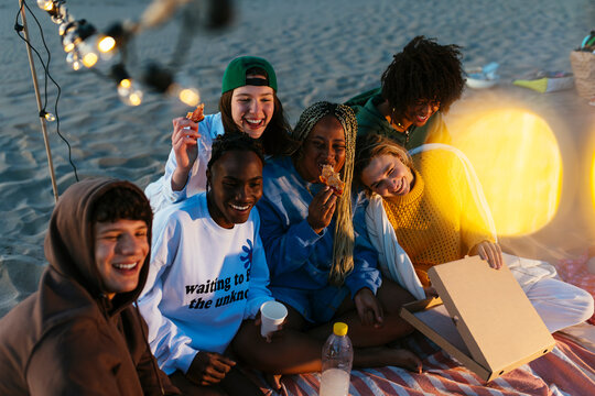Happy diverse friends eating pizza on beach at evening