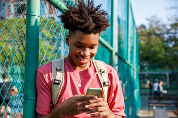 Smiling boy with backpack watching mobile phone while leaning on fence
