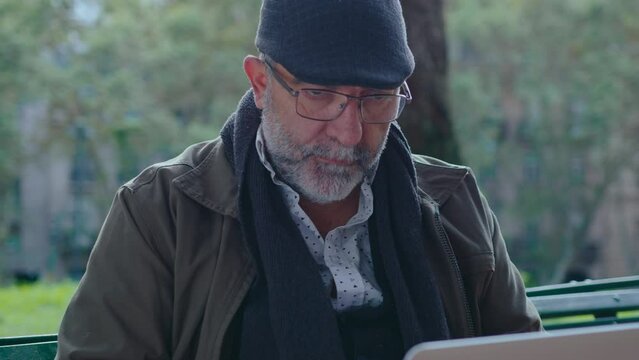 Senior man with gray beard wearing eyeglasses and cap sitting on bench in the park, chin stroking, thinking thoughtfully and using laptop outdoors