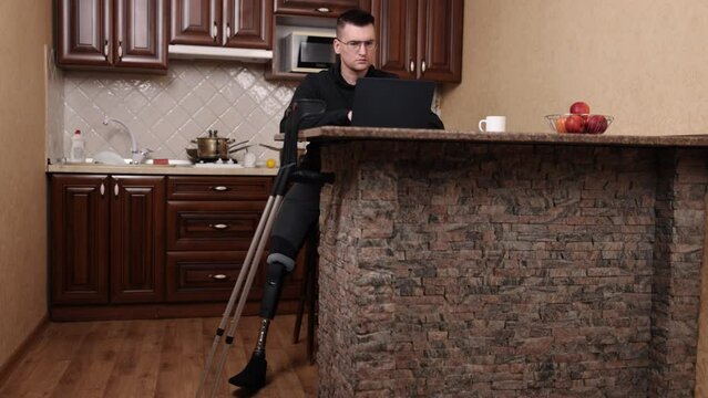 laptop work, impairment support, leg amputation. Man using leg prosthesis, wearing glasses, works on laptop with crutches at hand.