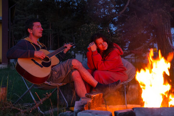 A man plays the guitar, a woman listens and sings along. A couple in love is sitting by the outdoor campfire in the courtyard of the house on camping chairs, a romantic evening