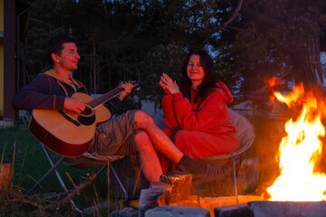 A man plays the guitar, a woman listens and sings along. A couple in love is sitting by the outdoor campfire in the courtyard of the house on camping chairs, a romantic evening