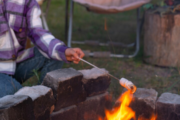 Fototapeta premium A girl in a plaid shirt roasts marshmallows on a fire in the yard of the house. Evening family get-together by the campfire, outdoor tourist chairs