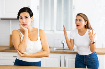 Fototapeta na wymiar Unhappy offended young Latina standing in kitchen at home, listening to her disgruntled female friend. Concept of problems in friendships