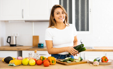 Obraz na płótnie Canvas Smiling young Hispanic woman standing in home kitchen, preparing fresh vegetable salad. Vegetarian diet concept